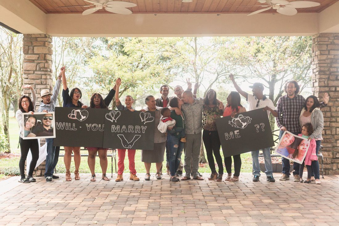 Family and friends hold signs and read will you marry me in a surprise proposal